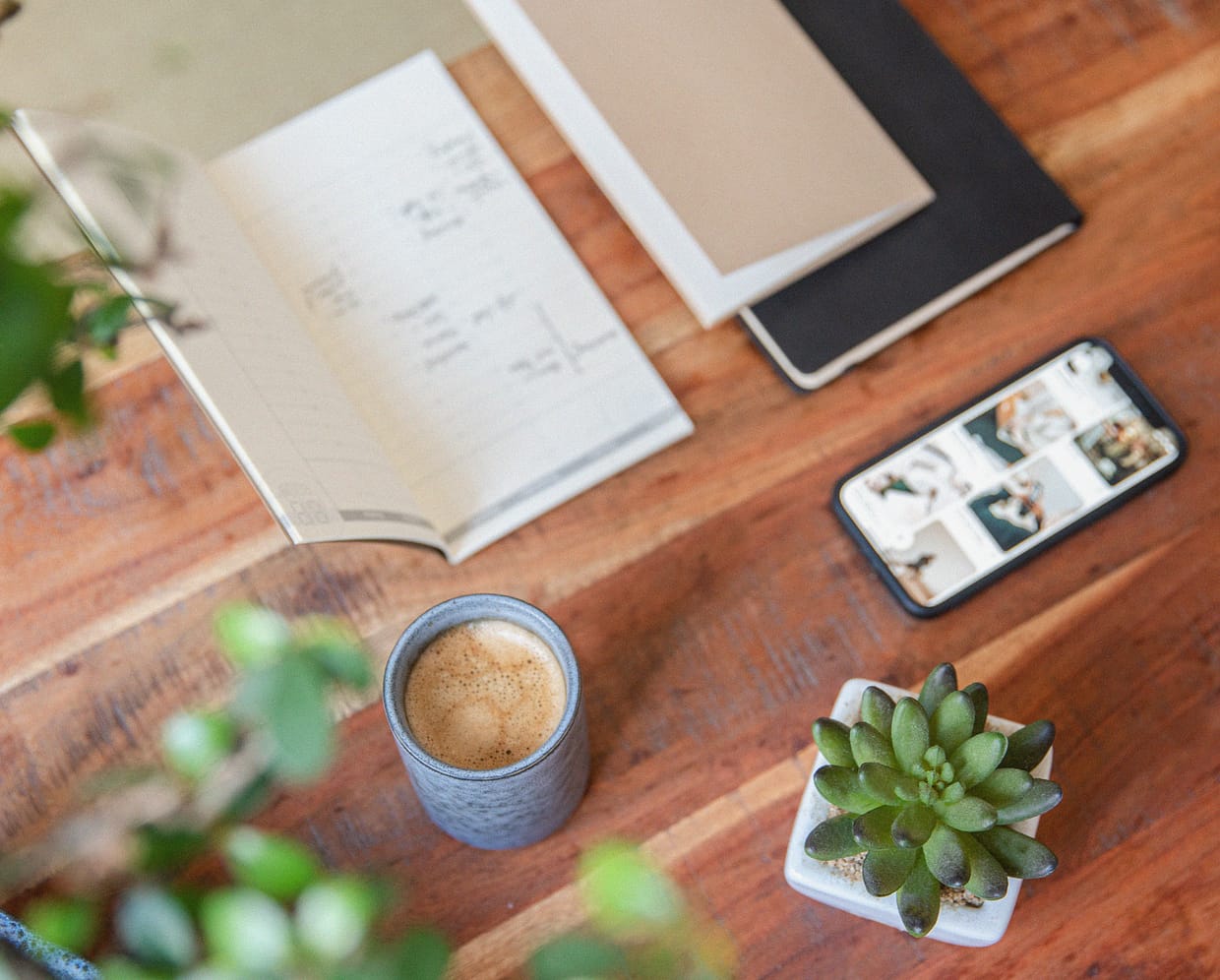 Auf einem Holztisch liegen diverse Büromaterialien, wie Notizhefte, Blume, Kaffeetasse und Handy