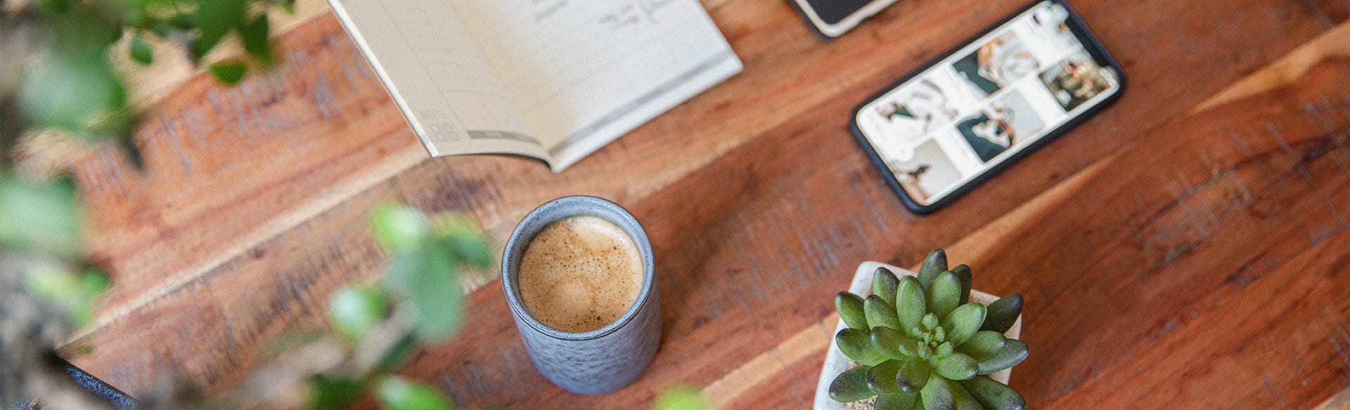 Auf einem Holztisch liegen diverse Büromaterialien, wie Notizhefte, Blume, Kaffeetasse und Handy